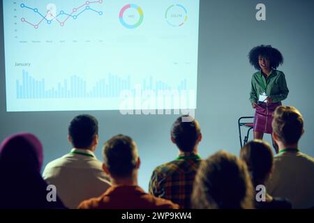 Selbstbewusste afroamerikanische Frau, Expertin mit Ständen vor dem Publikum, Vortrag mit Datendiagramm-Hintergrund im Konferenzraum. Stockfoto