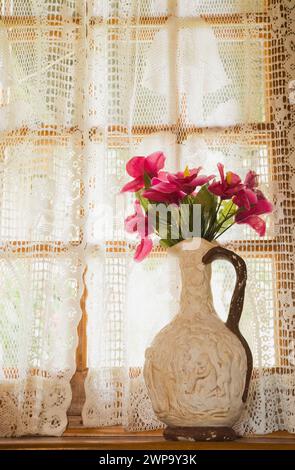 Keramikkkrug gefüllt mit roten Seidenblumen auf der Fensterbank in der alten rekonstruierten Blockhütte. Stockfoto