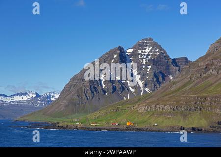 Zerklüftete Berge und orangefarbener Leuchtturm bei Skalanes entlang des Fjords Seyðisfjörður / Seydisfjoerdur im Sommer, Eastern Region / Austurland, Island Stockfoto