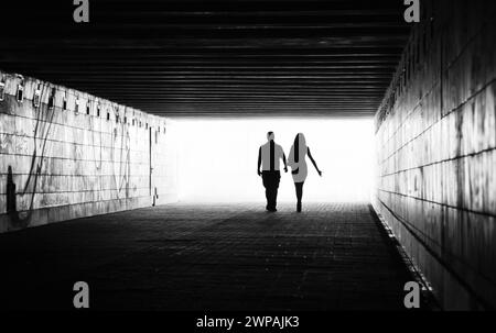 Silhouetten zweier Menschen in einem Tunnel - männliche und weibliche Figuren. Schwarzweißbild mit Licht von hinten. Ein Paar, das weggeht. Stockfoto