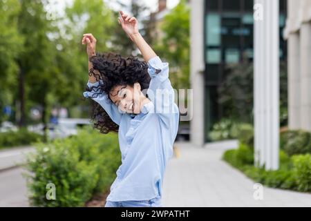 Eine überschwängliche junge Frau mit lockigen Haaren tanzt fröhlich auf einem urbanen Bürgersteig. Ihr lebendiger Ausdruck und ihre sorglose Bewegung vermitteln ein Gefühl der Freiheit und des Glücks in der städtischen Umgebung. Stockfoto