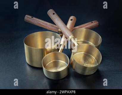 Satz Messbecher aus Messing mit Holzgriffen und hängendem Loch auf dunklem Hintergrund. Messbecher und Löffel aus Holz und Messing zum Messen von Trocken- und Messgeräten Stockfoto