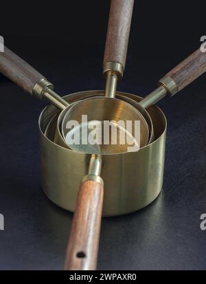 Satz Messbecher aus Messing mit Holzgriffen und hängendem Loch auf dunklem Hintergrund. Messbecher und Löffel aus Holz und Messing zum Messen von Trocken- und Messgeräten Stockfoto