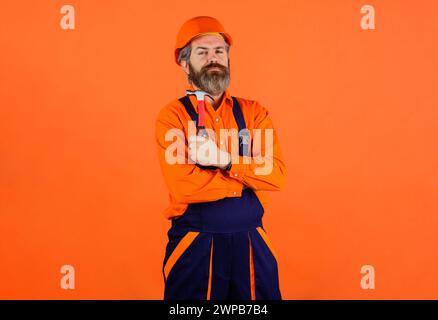 Bauherr im Bau Overall und Schutzhelm mit Hammer. Bärtiger Mann in Baumeister-Uniform und Helm mit Hammer. Vorarbeiter oder Zimmermann in Schutzkleidung Stockfoto