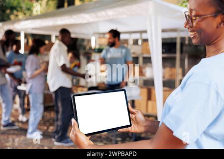 Das Bild zeigt eine freundliche afroamerikanische Dame, die ein Tablet mit Copyspace-Mockup-Vorlage für anpassbare Wohltätigkeitsnotizen trägt. Die lächelnde Frau greift das digitale Gerät horizontal mit einem isolierten weißen Bildschirm. Stockfoto