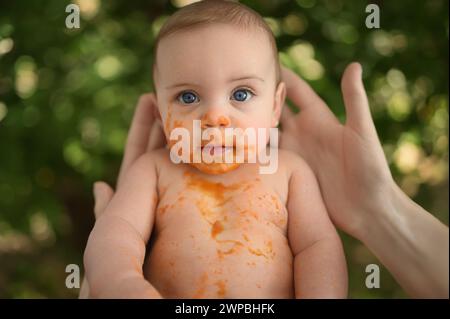 Niedliches entzückendes Baby lustiges schmutziges Baby mit Obst- oder Gemüsepüree auf seinem Gesicht. Erste kostenlose feste Speisen. Konzept für natürliche organische Inhaltsstoffe. Baby in Moms Händen Stockfoto