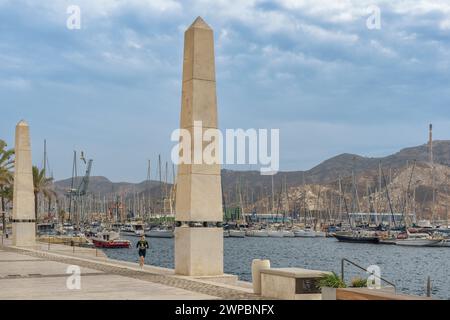 Foto einer Frau, die entlang der Esplanade des Hafens der Stadt Cartagena zwischen den beiden Säulen läuft, mit Booten, die im Hintergrund verankert sind, Murcia. Stockfoto