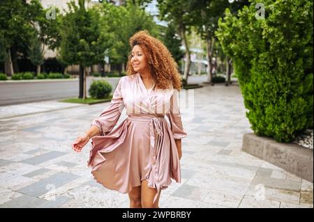 Eine junge, wunderschöne afroamerikanische Frau macht einen Spaziergang im öffentlichen Sommerpark. Eine glückliche Frau in Übergröße, die ein seidenrosa Kleid trägt, lächelt durch die Straßen der Stadt Stockfoto