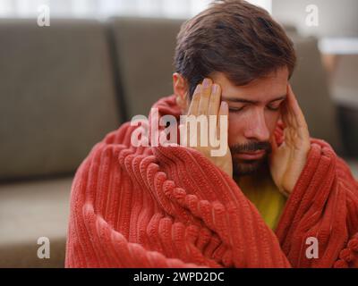 Herbst- und Wintersaison mit Niesen. Junger kaukasischer Mann mit Grippe fühlt sich kalt im Wohnzimmer zu Hause, hat Fieber und Kopfschmerzen und berührt seine Stirn, um seine Temperatur zu überprüfen. Stockfoto