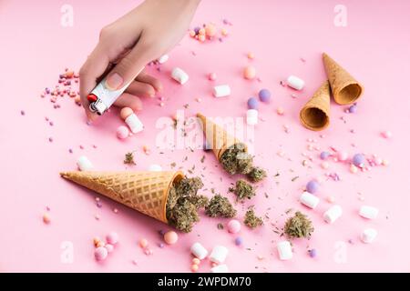 Trockene Knospen von medizinischem Marihuana liegen in Waffel-Eiskegeln, eine weibliche Hand hält ein Feuerzeug auf rosa Hintergrund. Es gibt Süßigkeiten und Marshmallows al Stockfoto