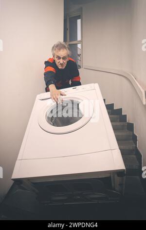 Ein älterer Mann, der die Waschmaschine auf dem Wagen im Gebäude bewegt Stockfoto