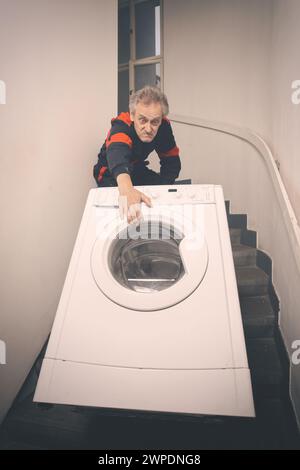 Ein älterer Mann, der die Waschmaschine auf dem Wagen im Gebäude bewegt Stockfoto