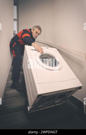 Ein älterer Mann, der die Waschmaschine auf dem Wagen im Gebäude bewegt Stockfoto