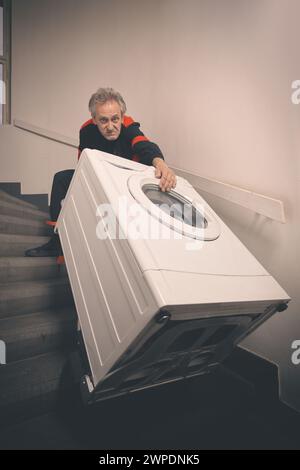 Ein älterer Mann, der die Waschmaschine auf dem Wagen im Gebäude bewegt Stockfoto