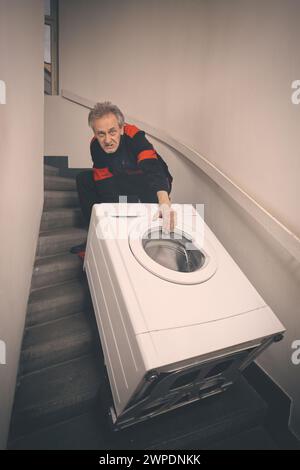 Ein älterer Mann, der die Waschmaschine auf dem Wagen im Gebäude bewegt Stockfoto