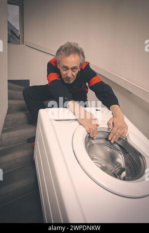 Ein älterer Mann, der die Waschmaschine auf dem Wagen im Gebäude bewegt Stockfoto