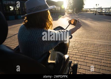 Frau Tourist auf einem Elektroroller mit vier Rädern auf einer Stadtstraße. Das Gesicht der Frau ist im Rückspiegel sichtbar. Stockfoto