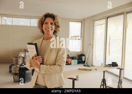 Glückliche Hausbesitzerin steht in ihrer Küche und hält Papiere für eine Hausrenovierung. Mit einem Lächeln schaut sie selbstbewusst in die Kamera und zeigt h Stockfoto