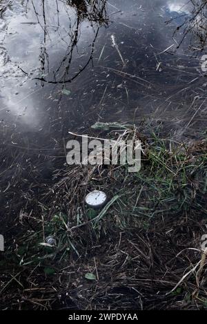 Eine alte silberne Taschenuhr, die im flachen Wasser am Rand eines Teichs liegt, Wolken spiegeln sich im Wasser. Stockfoto