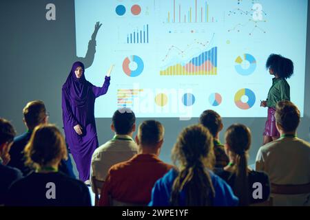 Der Sprecher im Hijab hebt die Hand, um den Punkt während der Präsentation hervorzuheben, während der Teilnehmer zur Bühne im Konferenzraum gehen. Stockfoto