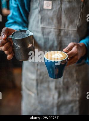 Barista mit Schürze gießt aufgeschäumte Milch in eine Kaffeetasse, wodurch ein flaches weiß entsteht Stockfoto