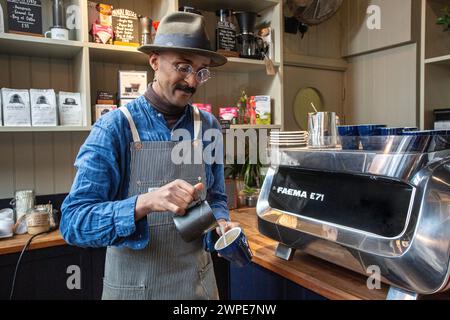 Barista mit Schürze gießt aufgeschäumte Milch in eine Kaffeetasse, wodurch ein flaches weiß entsteht Stockfoto