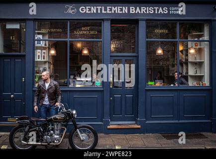 Ein junger Mann in Lederjacke macht eine Kaffeepause in einem Café in London mit einem klassischen Motorrad, das vor einem Café geparkt ist Stockfoto