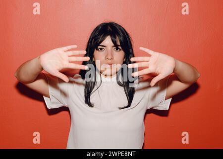 Porträt einer jungen lateinischen Frau, verzweifelt, verängstigt, mit den Händen davor, die auf rotem Hintergrund stehen, ernsthafte Gesten mit Gesicht und Händen machen. Stockfoto