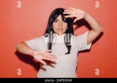 Porträt einer jungen lateinischen Frau, verzweifelt, verängstigt, mit den Händen davor, die auf rotem Hintergrund stehen, ernsthafte Gesten mit Gesicht und Händen machen. Stockfoto