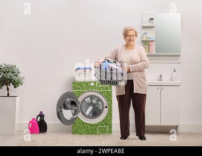 Porträt einer Reifen Frau in voller Länge, die einen Wäschekorb mit Kleidung in einem Badezimmer mit umweltfreundlicher Waschmaschine hält Stockfoto
