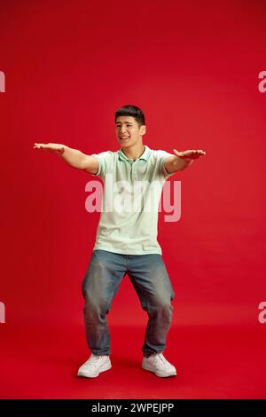 Ganzkörperfoto eines lustigen, fröhlichen, jungen asiatischen Mannes in lässiger Kleidung, der mit Freude in Bewegung vor rotem Studio-Hintergrund tanzt. Stockfoto