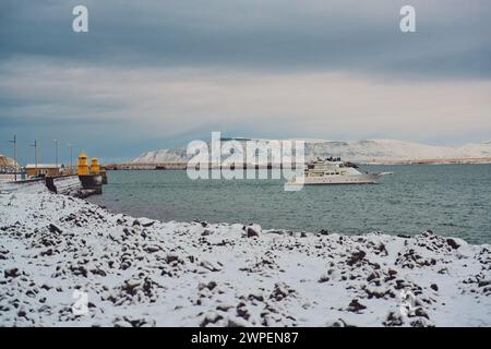 Winterlandschaft mit schneebedeckter Küste und Boot auf kaltem, blauem Wasser mit Bergen im Hintergrund. Lage: Reykjavik Island. Stockfoto