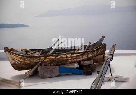 Ein altes Holzboot, das die Spuren vieler Reisen trägt, befindet sich von einem Dach in Santorin mit Blick auf die weite Ägäis. Stockfoto