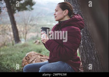 Junge Frau, die in der Natur des Waldes chillt, eine Tasse heißes Getränk hält, träumerisch in die Ferne blickt. Menschen. Ländlicher Lebensstil. Aktiv und hea Stockfoto