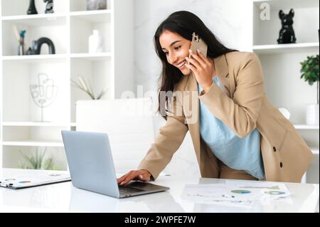 Telefonieren. Eine geschäftige, hübsche indische oder arabische Geschäftsfrau, Firmenangestellte, ceo, steht neben einem Tisch im Büro, spricht per Smartphone, führt Gespräche mit Partner oder Kunden, benutzt Laptop, lächelt Stockfoto