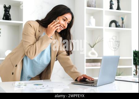 Smartphone-Konversation. Indische oder arabische Geschäftsfrau, Unternehmensleiter, ceo, steht neben einem Tisch im Büro, spricht per Smartphone, verhandelt mit Partner oder Kunden, benutzt Laptop, lächelt Stockfoto
