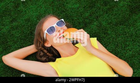 Porträt einer fröhlich lächelnden jungen Frau mit Burger-Fast-Food auf grünem Gras im Sommerpark Stockfoto