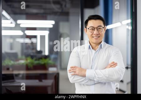 Ein professioneller asiatischer Geschäftsmann steht selbstbewusst mit gekreuzten Armen und lächelt in einer modernen Büroumgebung. Stockfoto