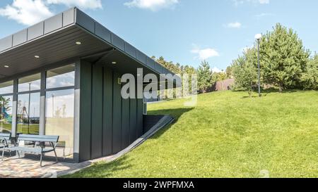 Modernes Gästehaus mit stehender Nahtfassade, Panoramaverglasung und großem Flachdach-Überhang im Park. Minimalistischer architektonischer Stil des öffentlichen Gebäudes Stockfoto