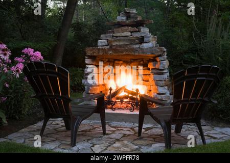 Zwei Adirondack-Stühle vor dem holzbrennenden Natursteinkamin auf der Pflasterterrasse im Garten im Garten im Garten im Sommer in der Abenddämmerung. Stockfoto