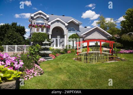 Graue Ziegelsteine mit weißer Zierleiste, zweistöckiges Haus und roter Holzsteg über Teich und Rand mit Pagode, rosa und rote Impatiens, Petunia-Blumen. Stockfoto