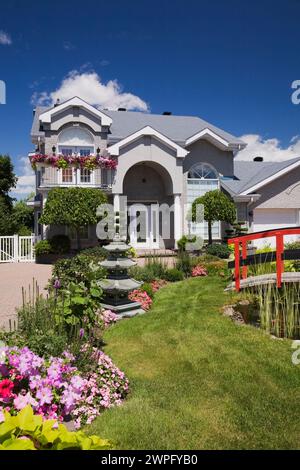 Graue Ziegelsteine mit weißer Zierleiste, zweistöckiges Haus und roter Holzsteg über Teich und Rand mit Pagode, rosa und rote Impatiens, Petunia-Blumen. Stockfoto