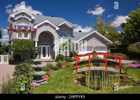 Graue Ziegelsteine mit weißer Zierleiste, zweistöckiges Haus und roter Holzsteg über Teich und Rand mit Pagode, rosa und rote Impatiens, Petunia-Blumen. Stockfoto