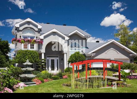 Graue Ziegelsteine mit weißer Zierleiste, zweistöckiges Haus und roter Holzsteg über Teich und Rand mit Pagode, rosa und rote Impatiens, Petunia-Blumen. Stockfoto