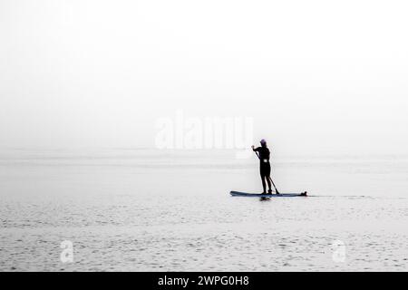 Silhouette einer friedlichen Solo-Frau auf einem Paddleboard, die alleine im dichten, unheimlichen Nebel auf einem See oder Ozean erkundet Stockfoto