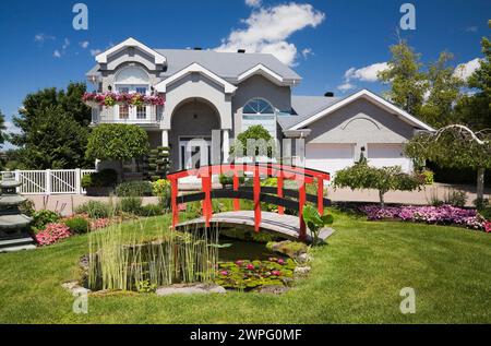 Graue Ziegelsteine mit weißer Zierleiste, zweistöckiges Haus und roter Holzsteg über Teich und Rand mit Pagode, rosa und rote Impatiens, Petunia-Blumen. Stockfoto