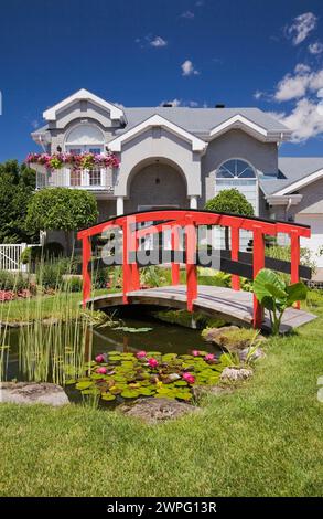 Graue Ziegelsteine mit weißer Zierleiste, zweistöckiges Haus und roter Holzsteg über Teich und Rand mit Pagode, rosa und rote Impatiens, Petunia-Blumen. Stockfoto