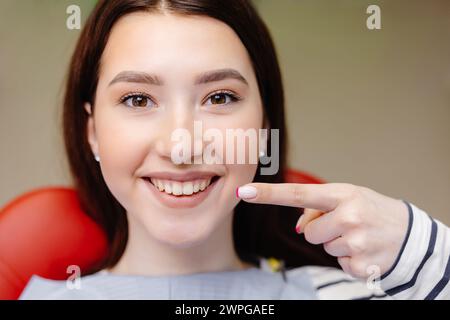 Zugeschnittenes Nahfoto eines jungen lächelnden Mädchens, bedeckt mit Serviette, Zeigefinger auf gesundes zahniges Lächeln, das auf dem Zahnarztstuhl sitzt. Gesundheit Stockfoto