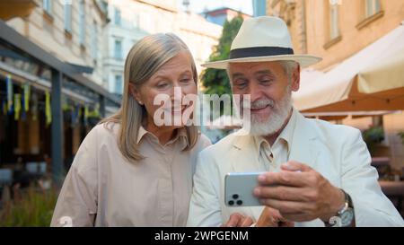 Glückliches lächelndes kaukasisches älteres Ehepaar zu Fuß Selfie machen Lachen sprechendes Gadget Handy zusammen Stadtstraße draußen. Alte Frau Stockfoto