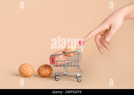 Weibliche Hand mit kleinem Einkaufswagen und Ostereiern auf beigefarbenem Hintergrund Stockfoto